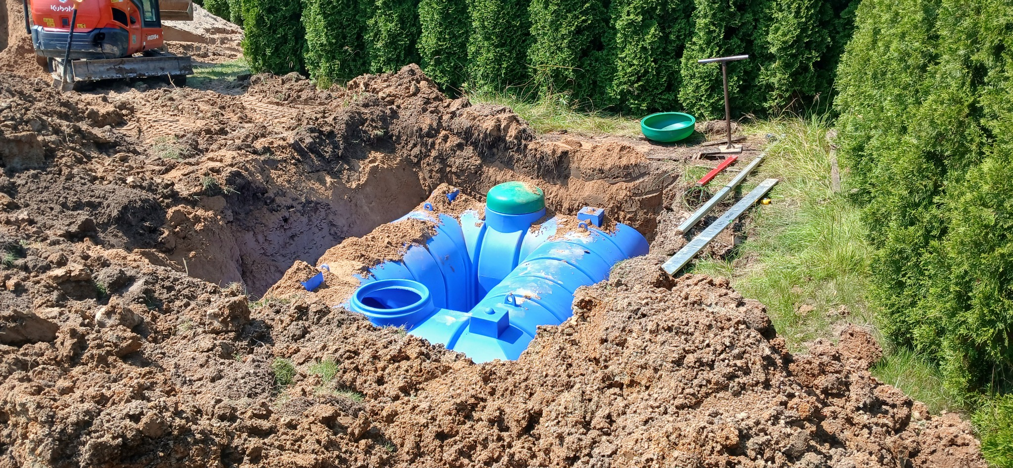 Montaż przydomowej oczyszczalni ścieków: niebieski zbiornik z zieloną pokrywą częściowo zasypany ziemią w wykopie, widoczny minikoparka Kubota, poziomice i inne narzędzia, zielone tuje w tle.