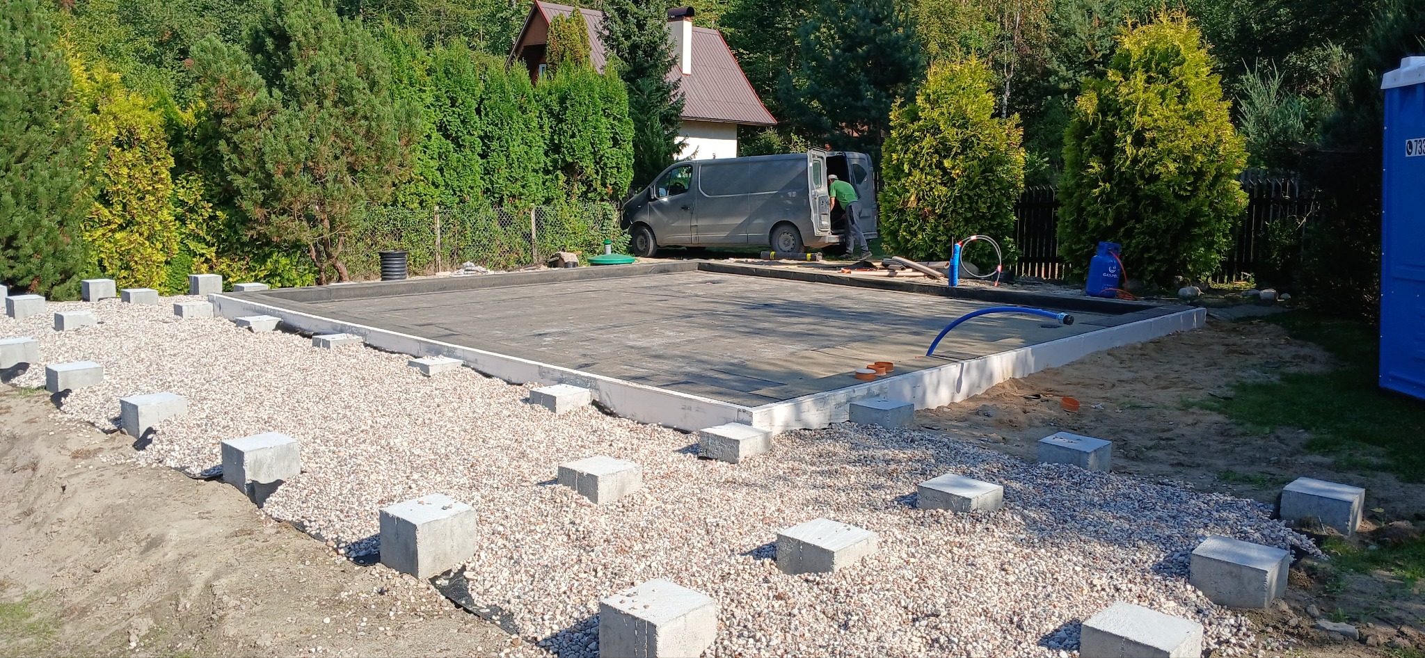 Przygotowany teren pod budowę, z widocznymi betonowymi bloczkami na żwirowym podłożu, izolacją i pracownikiem przy samochodzie dostawczym w tle.