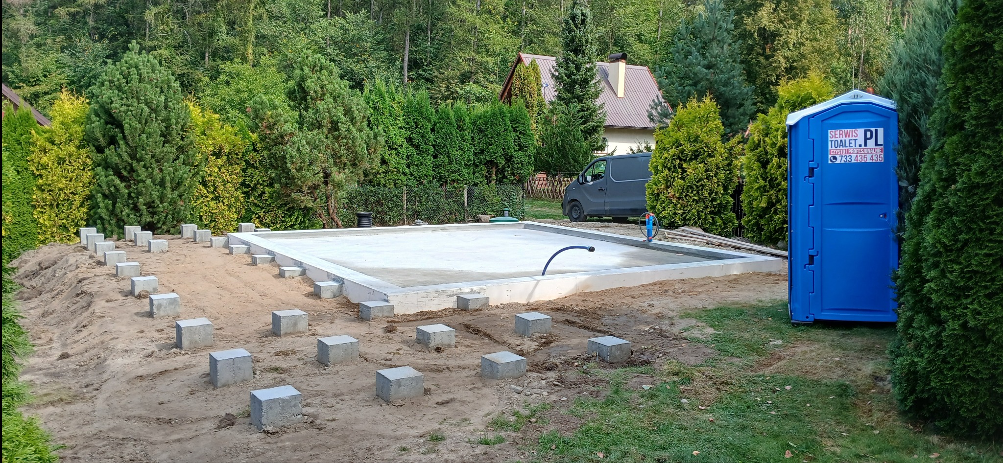 Wylany fundament pod budowę domu z widocznymi betonowymi słupkami na obrzeżach, niebieska przenośna toaleta Toalet.pl z boku, w tle zielony ogród i szary samochód dostawczy.
