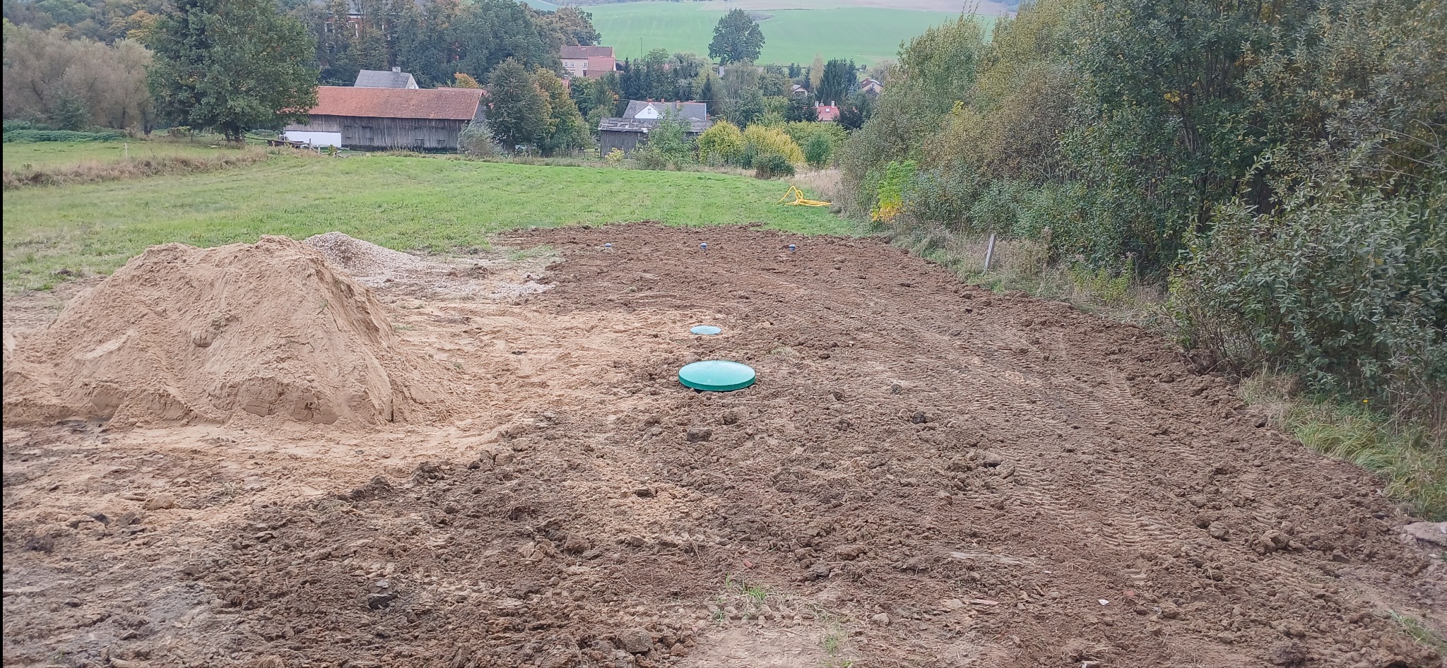 Widok na plac budowy z pryzmą piasku i trzema zielonymi pokrywami włazów, prawdopodobnie od przydomowej oczyszczalni ścieków, na tle wiejskich zabudowań i drzew.