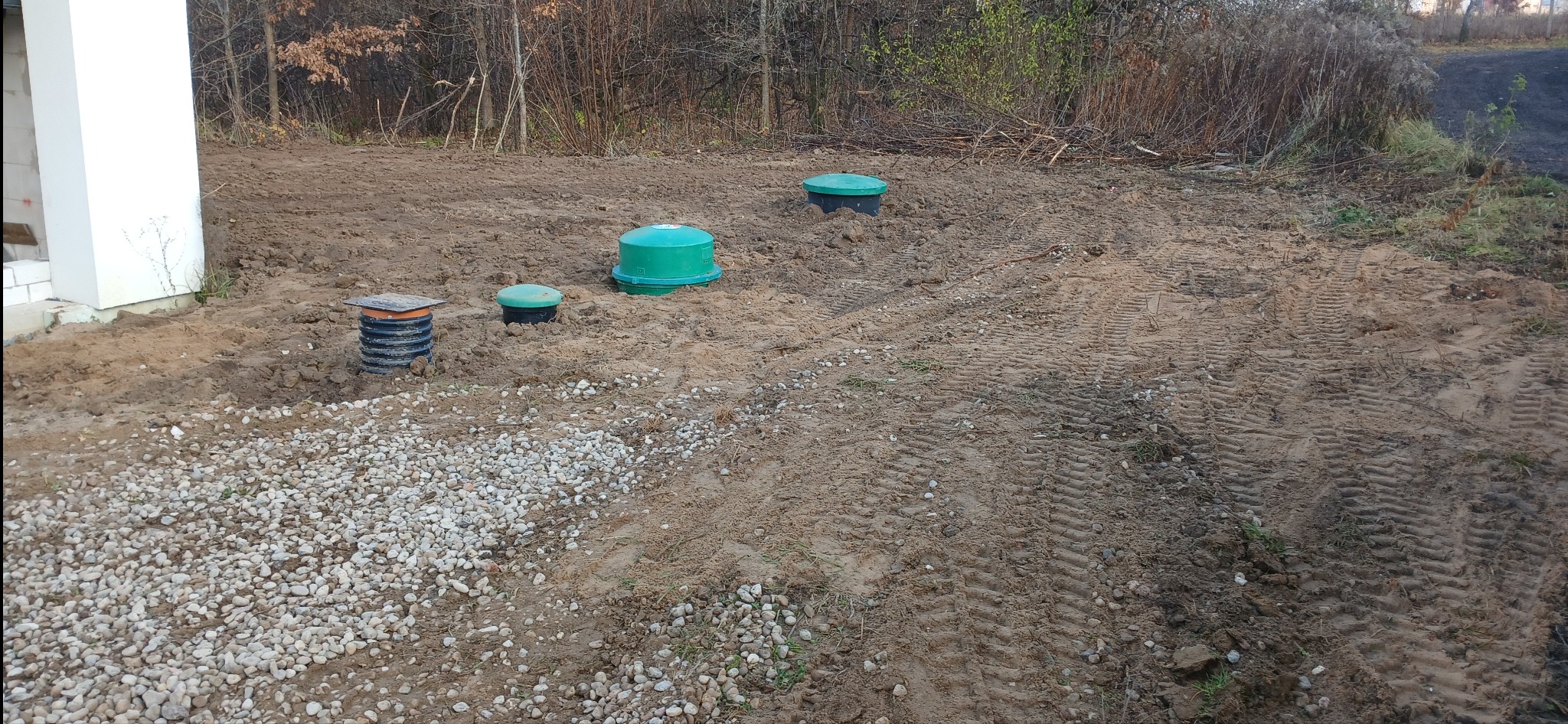 Instalacja przydomowej oczyszczalni ścieków z widocznymi włazami i drenażem żwirowym, świeżo wyrównany teren z śladami opon, obok fundamentu budynku.