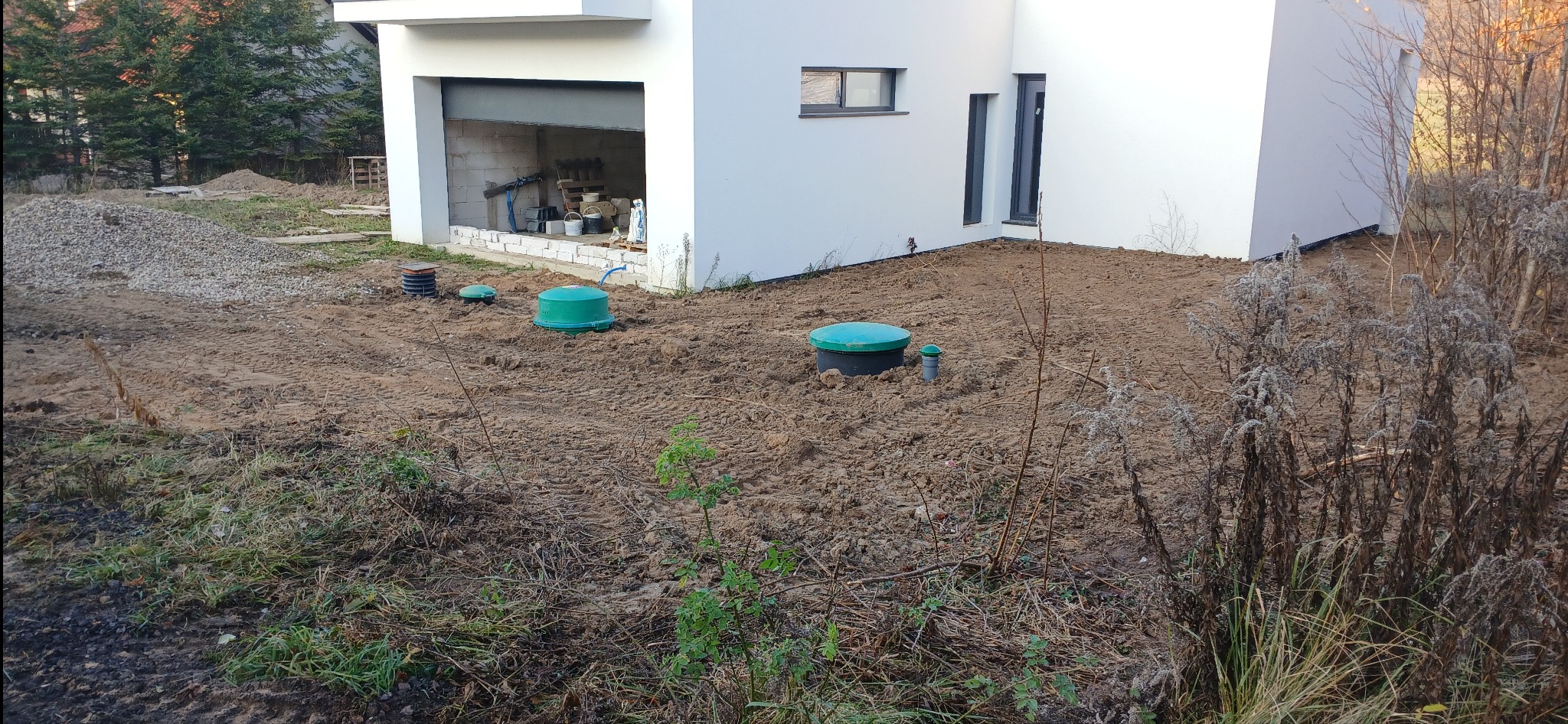 Teren po wyrównaniu podłoża z widocznymi elementami instalacji przydomowej oczyszczalni ścieków, z fragmentem budynku w tle i stertą gruzu.