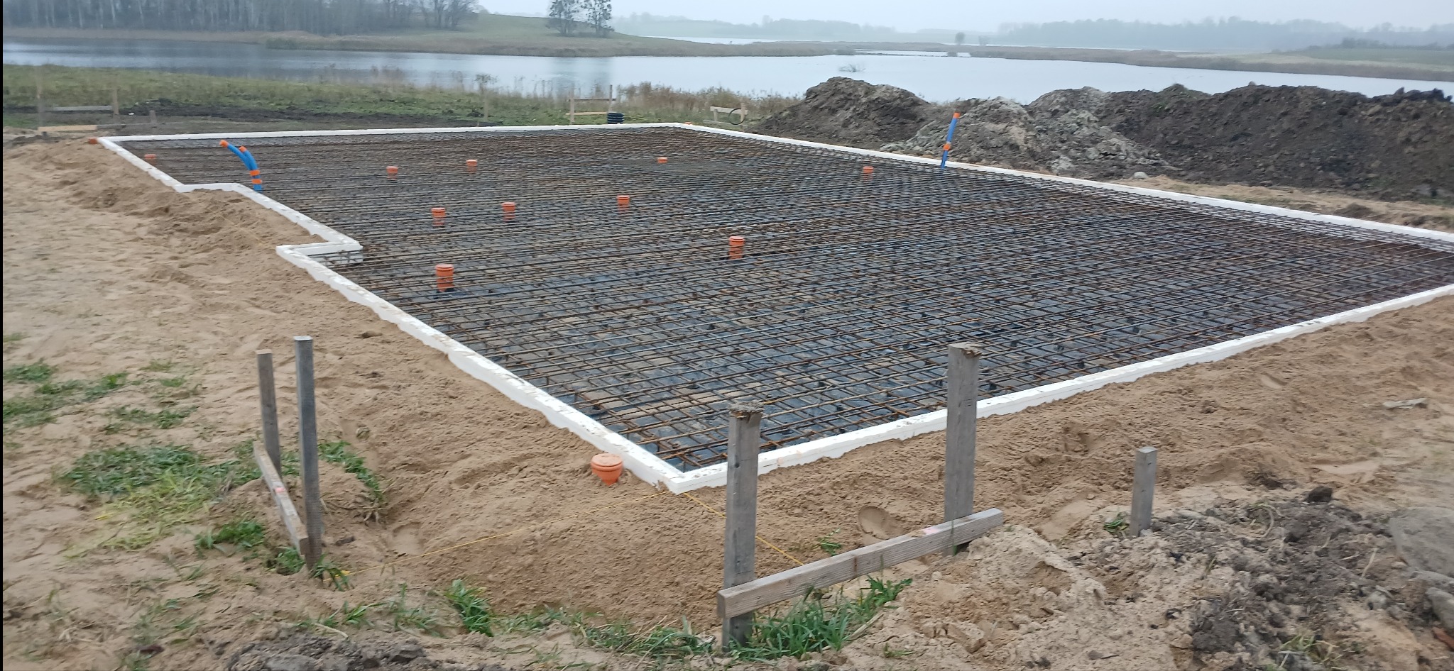 Zbrojenie fundamentu płyty, otoczone białym styropianem, z pomarańczowymi rurami wentylacyjnymi, przygotowane do zalania betonem, na piaszczystym terenie z widokiem na jezioro w tle.