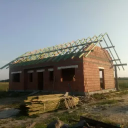 Dom murowany w stanie surowym otwartym z widoczną więźbą dachową, stos drewna na pierwszym planie, teren niezabudowany w tle.