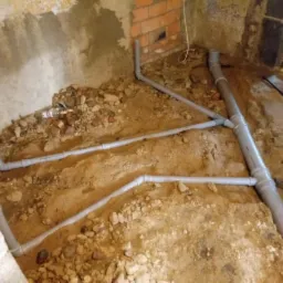 Szare rury kanalizacyjne ułożone w wykopie w ziemi, widoczne fragmenty fundamentów i ściany z cegły, instalacja pod posadzką w trakcie budowy.