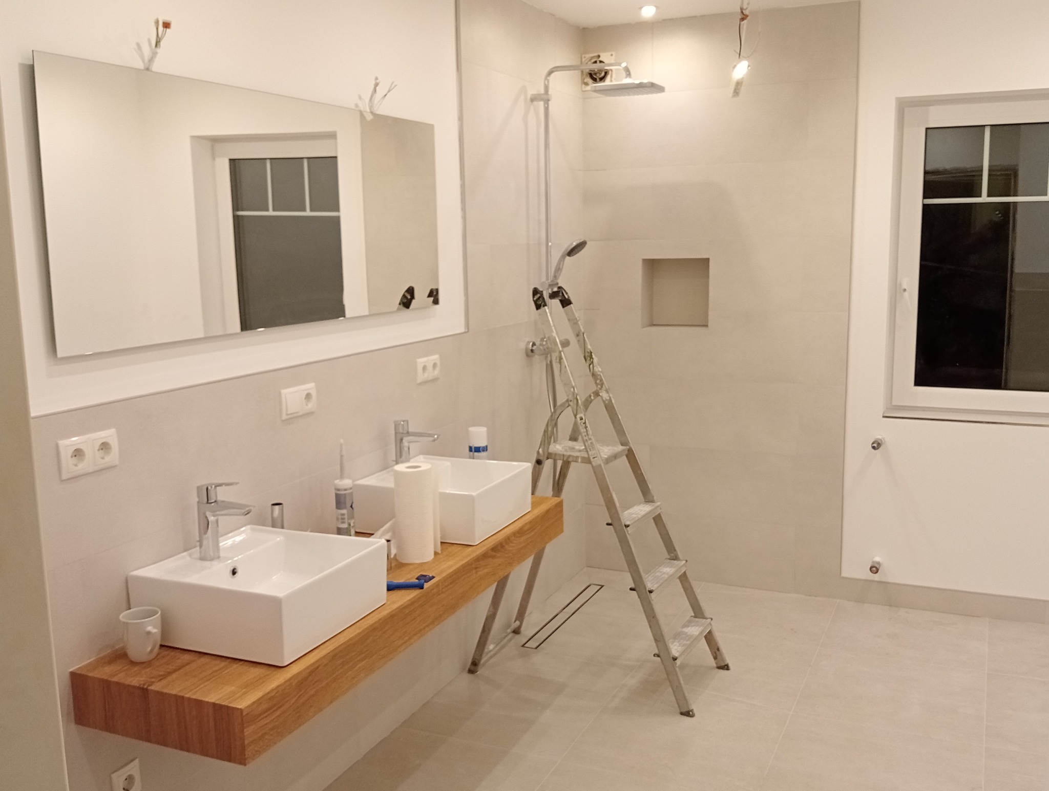 Nowoczesna łazienka w trakcie wykończenia: dwie białe umywalki na drewnianym blacie, lustro, kabina prysznicowa z deszczownicą, drabina, białe płytki na ścianach i podłodze, okno z białymi ramami.
