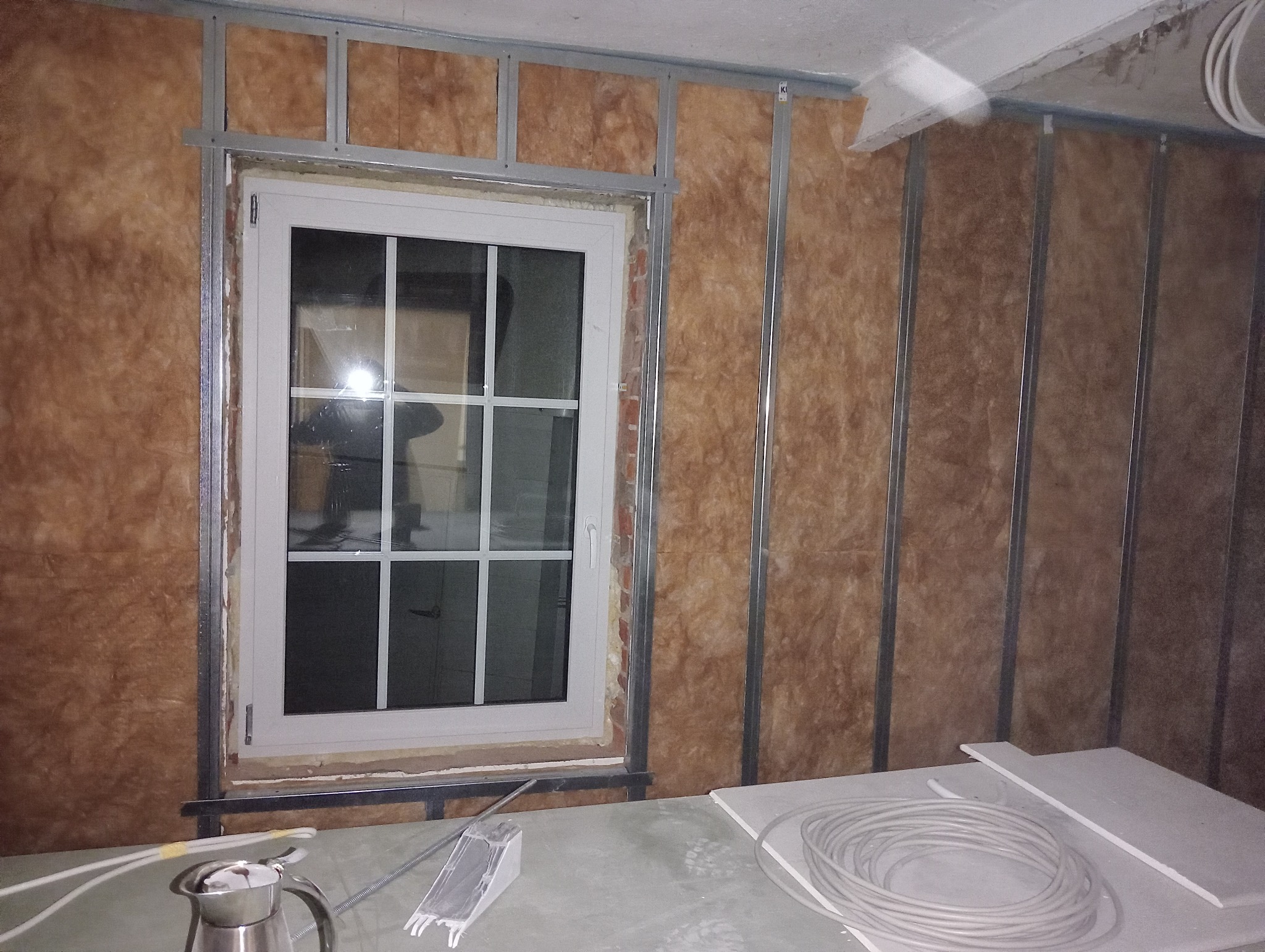 Wnętrze w trakcie ocieplania, widoczna metalowa konstrukcja, izolacja z wełny mineralnej, okno PCV i materiały budowlane na pierwszym planie.