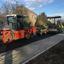 Walec HAMM i rozściełacz Volvo pracują na nowo wyasfaltowanej drodze w słoneczny dzień. Widoczni robotnicy w odblaskowych kamizelkach i budynki w tle.