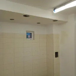 Wnętrze łazienki w trakcie remontu, widoczne beżowe płytki na ścianach, otwory na oświetlenie w suficie podwieszanym, wystające kable i częściowo pomalowane ściany.