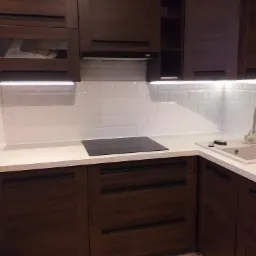 Nowoczesna kuchnia z ciemnymi drewnianymi szafkami, białym blatem i białymi płytkami ściennymi, oświetlona listwami LED pod szafkami górnymi.