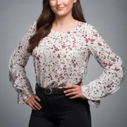 Portret kobiety w kwiecistej bluzce z rękawami z falbankami i czarnych spodniach, pozującej na szarym tle.