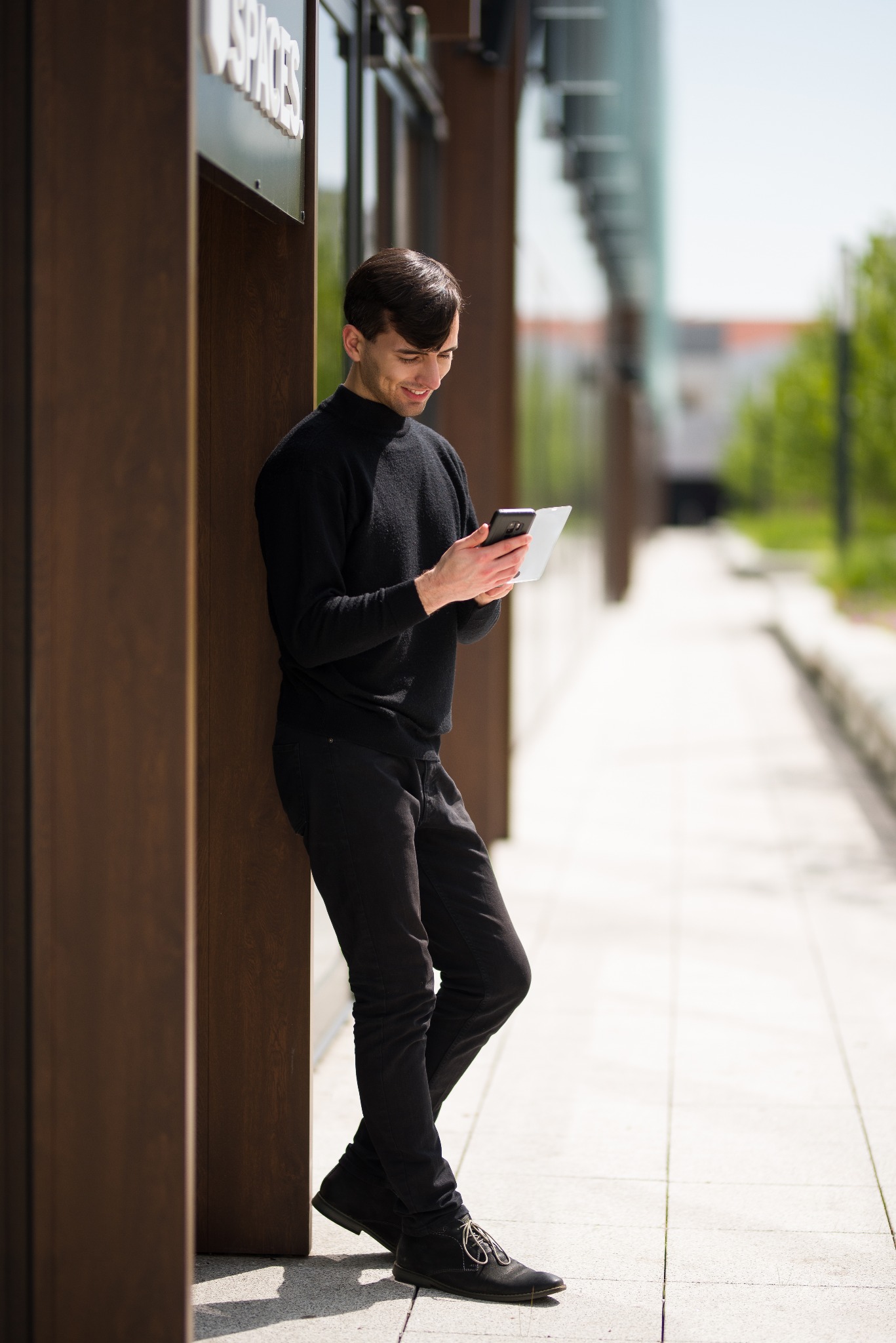 Młody mężczyzna w czarnym ubraniu oparty o brązowy filar budynku, przeglądający tablet na zewnątrz.