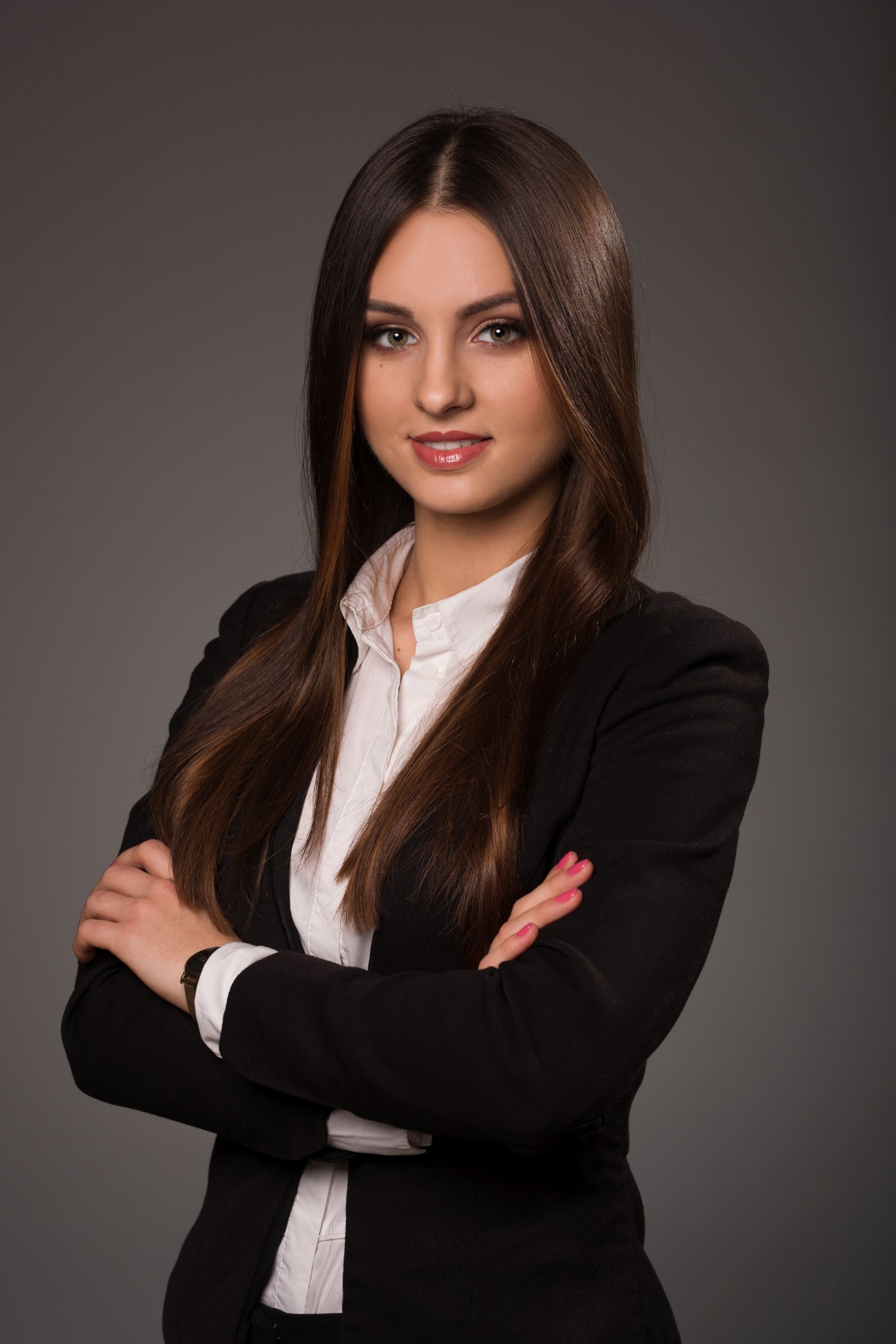 Portret kobiety w czarnej marynarce i białej koszuli, z założonymi rękami, na szarym tle.