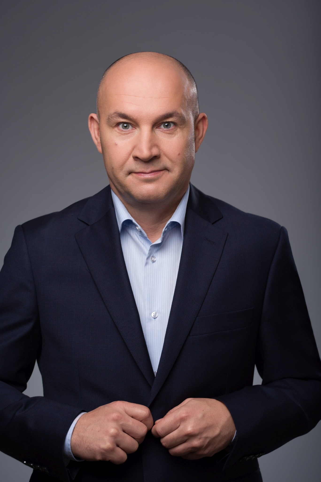 Portret biznesowy mężczyzny w granatowym garniturze i błękitnej koszuli na szarym tle, zapinającego guzik marynarki.