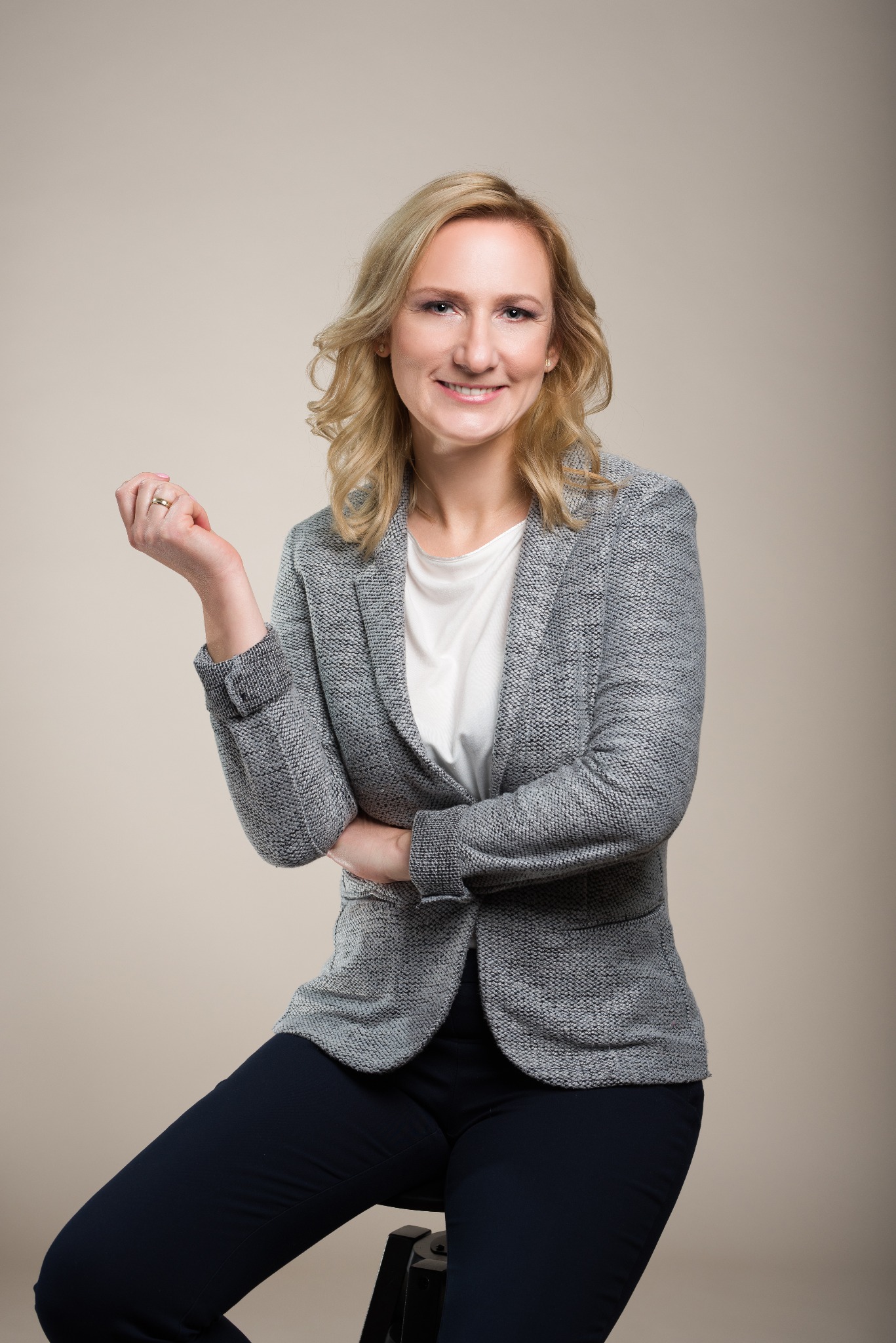 Portret uśmiechniętej blondynki w szarym żakiecie i białej koszuli, siedzącej na krześle na neutralnym tle.