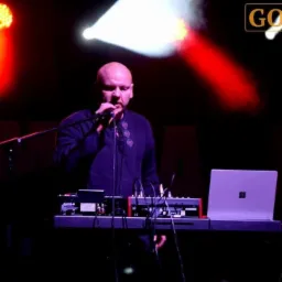 Wokalista z mikrofonem i klawiszami, oświetlony czerwonym i białym światłem scenicznym, obok laptop