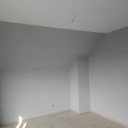 Pomalowane na szaro wnętrze pokoju na poddaszu z drewnianymi listwami przypodłogowymi i grzejnikiem przy ścianie