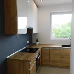 Nowoczesna kuchnia z białymi szafkami górnymi i drewnopodobnymi szafkami dolnymi, czarnym blatem i płytą grzewczą, szara ściana z dekoracyjnym tynkiem, okno z widokiem na zieleń, minimalistyczne...