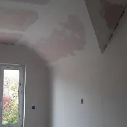 Pokój w trakcie remontu: fragment sufitu z widocznymi plamami po gruntowaniu, ściany przygotowane pod malowanie, okno z widokiem na zieleń za nim, gniazdko elektryczne na ścianie.