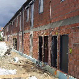 Wznoszenie szeregu budynków z czerwonej cegły, widoczne okna i drzwi z folią ochronną, teren budowy z paletami i gruzem, oznaczenia literowe na ścianach.