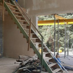 NVK Group Sp. z o.o. - Wnętrze w trakcie budowy domu z drewnianymi schodami, częściowo otynkowanymi ścianami i widoczną konstrukcją dachu z oknem dachowym. Na podłodze leżą materiały budowlane i narzędzia.