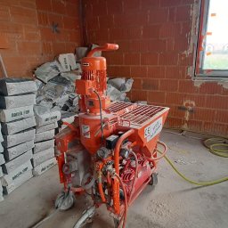 NVK Group Sp. z o.o. - Pomarańczowa maszyna do nakładania zaprawy stoi na budowie, obok sterta worków z zaprawą, ściany z czerwonej cegły, widoczne przewody i okno.