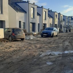 NVK Group Sp. z o.o. - Rząd nowo wybudowanych, surowych domów jednorodzinnych z szarymi dachami i oknami zabezpieczonymi folią, stojących wzdłuż błotnistej drogi z zaparkowanymi samochodami.