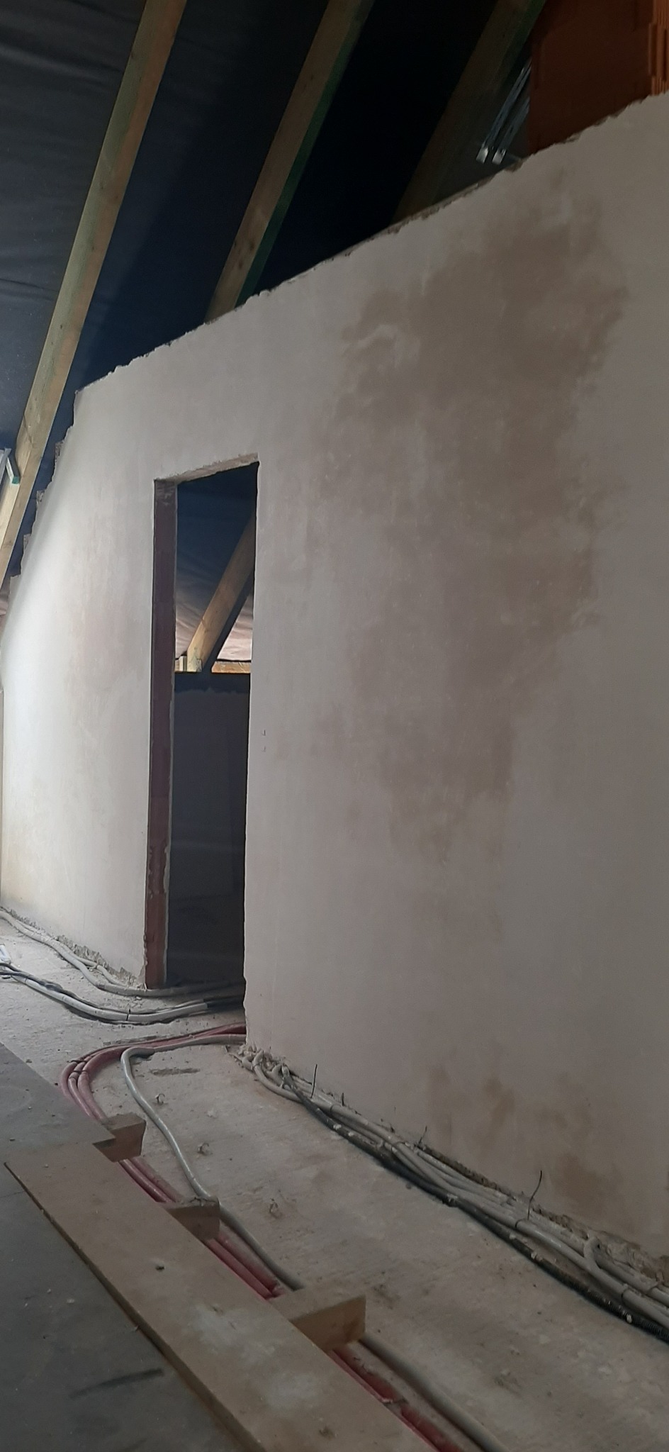 Ściana poddasza po nałożeniu świeżej warstwy szarej zaprawy, widoczne drewniane elementy konstrukcyjne dachu i instalacje elektryczne biegnące wzdłuż podłogi, otwór drzwiowy w stanie surowym.