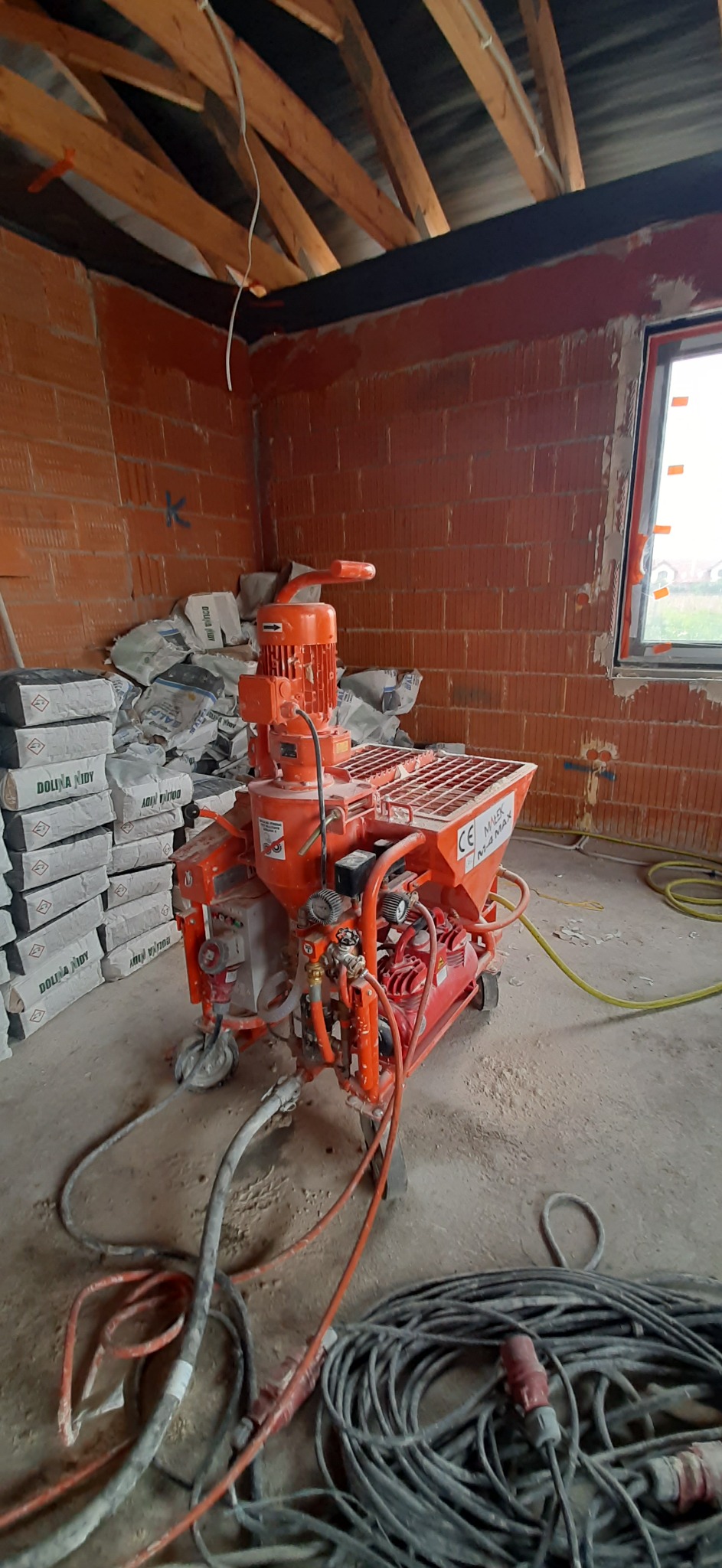 Pomarańczowa maszyna do nakładania zaprawy stoi na budowie, obok sterta worków z zaprawą, ściany z czerwonej cegły, widoczne przewody i okno.