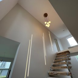DOM4YOU - Wysokie wnętrze z nowoczesnym oświetleniem: pionowe listwy LED na ścianie klatki schodowej i wisząca lampa z kulistymi kloszami. Schody zabezpieczone kartonami, widok na okno i fragment budynku...