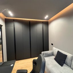 DOM4YOU - Nowoczesny pokój z podświetlanym sufitem, czarną zabudową szafy wnękowej z ciemnymi listwami, szarą sofą z czarną poduszką i fotelem gamingowym.