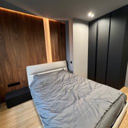 DOM4YOU - Nowoczesna sypialnia z łóżkiem w szarym odcieniu, drewnianą ścianą z podświetleniem LED i czarną szafą wnękową.