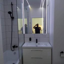 DOM4YOU - Nowoczesna łazienka z białymi płytkami, czarną armaturą i podświetlanym lustrem, odbicie osoby robiącej zdjęcie.