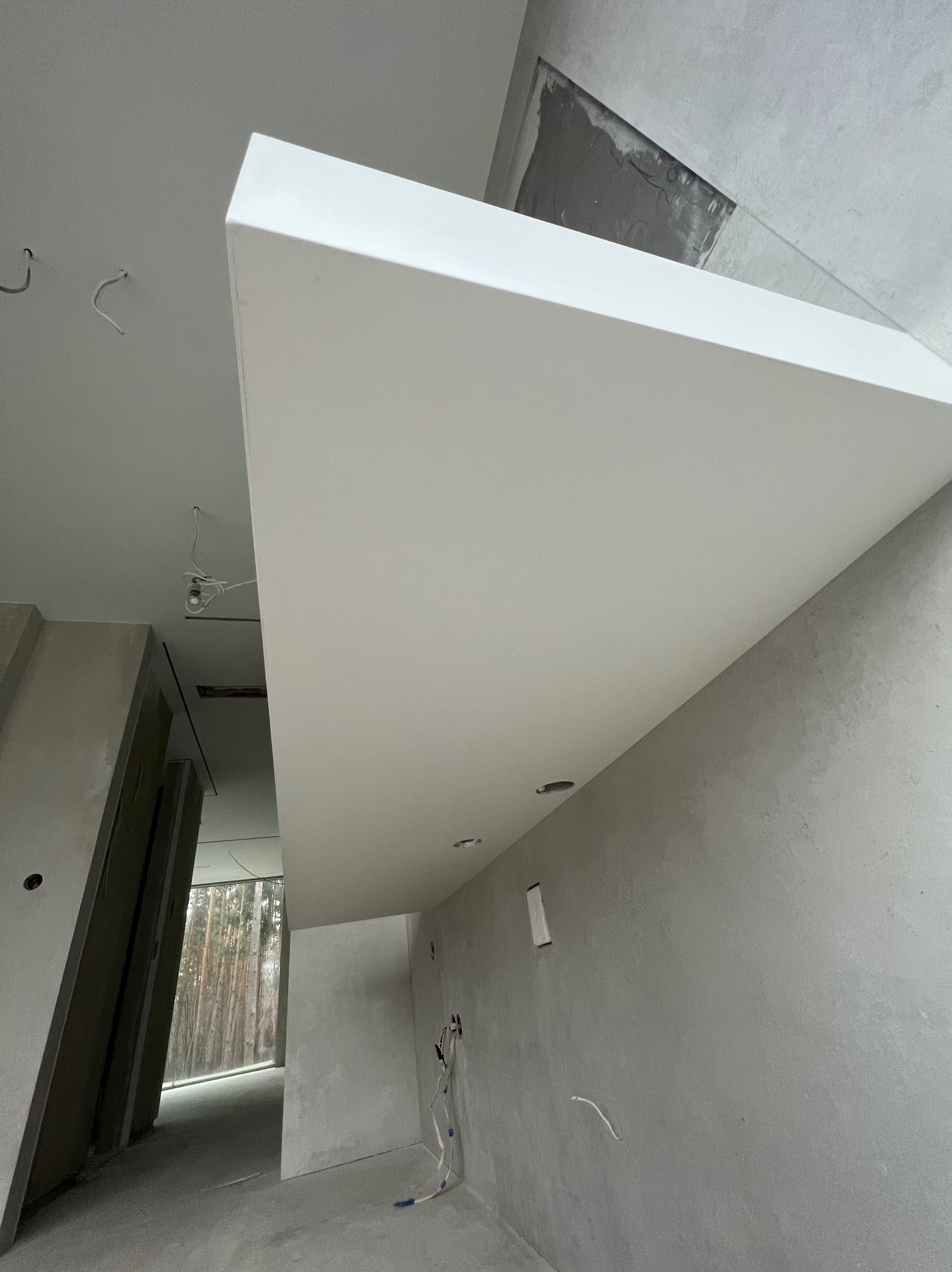 Surowy stan wnętrza z widoczną konstrukcją podwieszanego sufitu z płyt kartonowo-gipsowych, nieotynkowane ściany, instalacje elektryczne w trakcie montażu, widok na okno z lasem w tle.