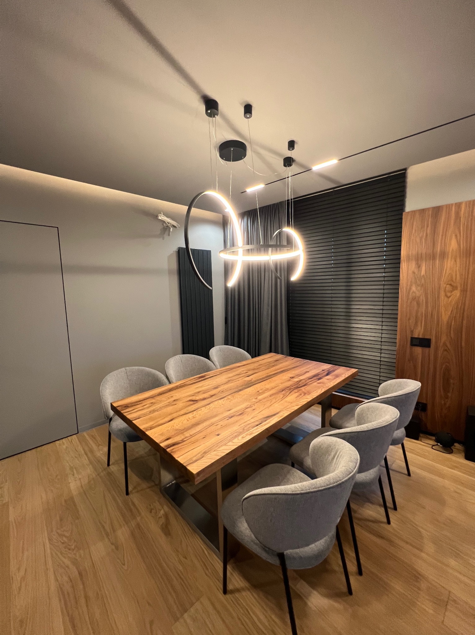 Nowoczesna jadalnia z drewnianym stołem, szarymi krzesłami i designerskimi lampami wiszącymi w kształcie pierścieni, uzupełniona o żaluzje i drewnianą okładzinę ściany.