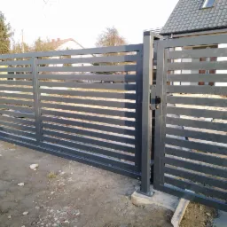 Nowoczesna, przesuwna, metalowa brama wjazdowa z poziomymi szczeblami i furtką, świeżo zamontowana na posesji z widocznym surowym podłożem.