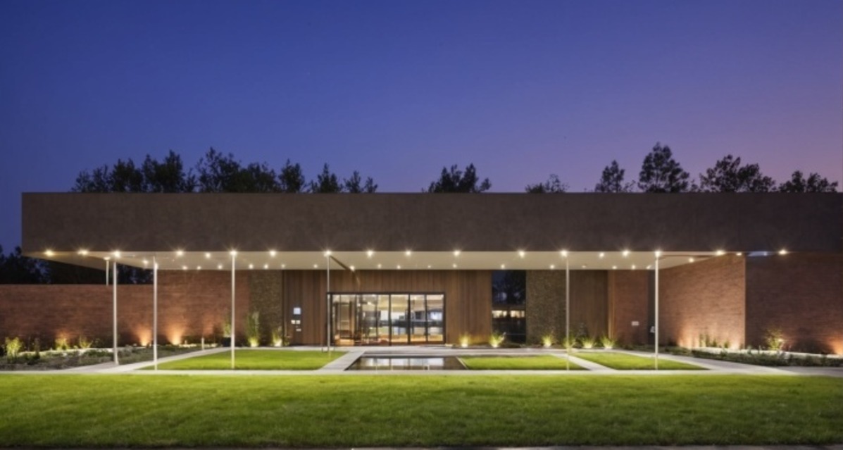 Nowoczesny budynek usługowy z płaskim dachem i oświetlonym wejściem, otoczony zielonym trawnikiem i niską roślinnością, sfotografowany o zmierzchu.