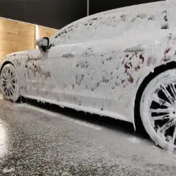 Czerwony samochód pokryty gęstą pianą podczas mycia w myjni, widok z boku na felgi i karoserię.