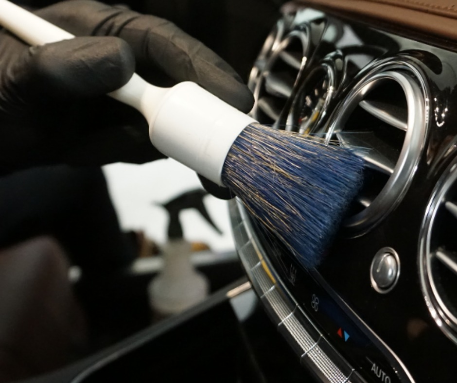 Szczegółowe czyszczenie szczotką z niebieskim włosiem kratki wentylacyjnej w luksusowym samochodzie z elementami chromowanymi, widoczne dłonie w czarnych rękawiczkach.