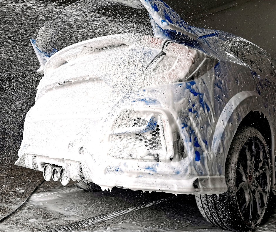 Niebieski samochód sportowy pokryty grubą warstwą piany podczas mycia, widok z tyłu na potrójny wydech i czarne felgi.