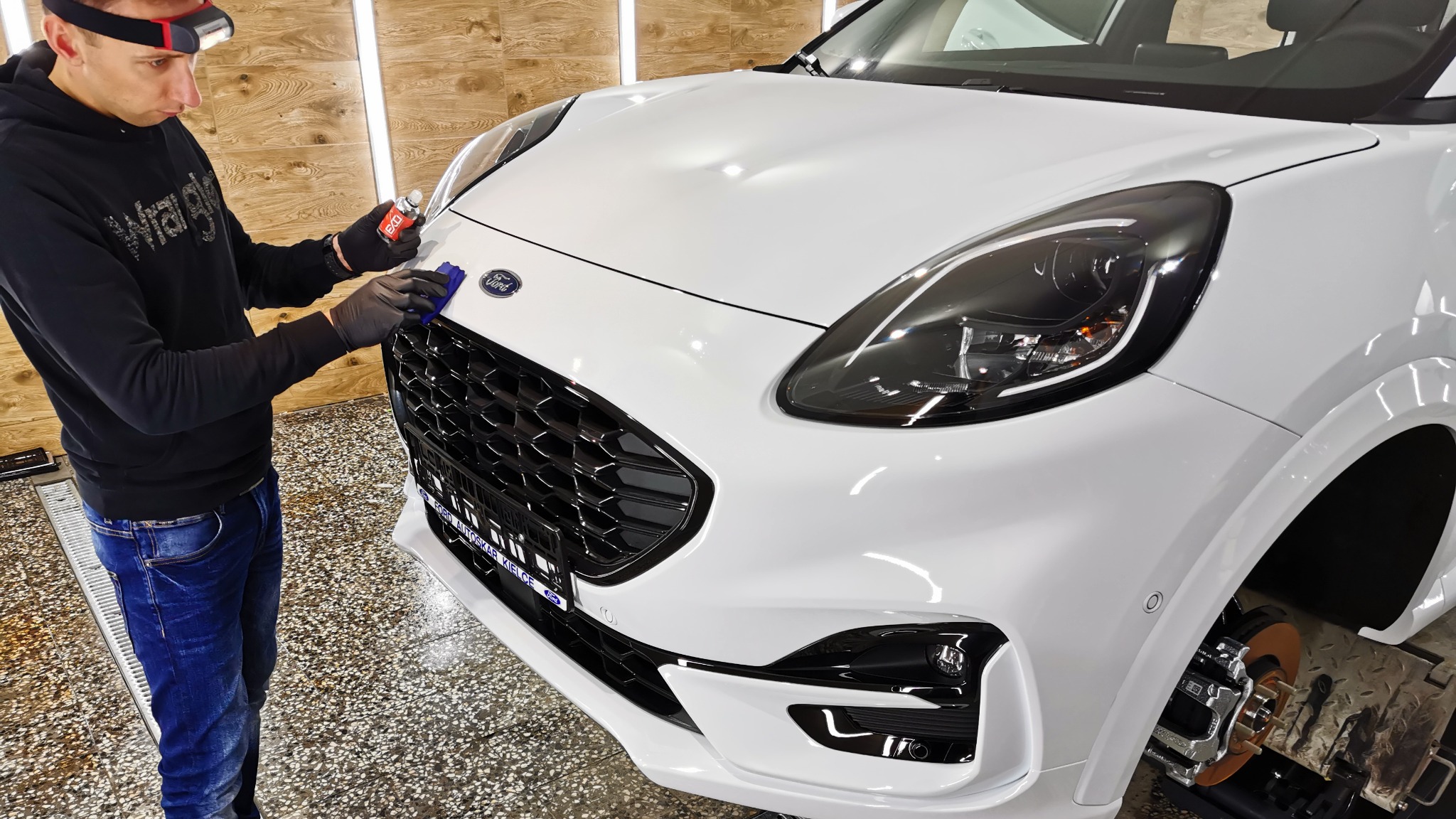 Pracownik w czarnych rękawicach aplikuje preparat na przedni grill białego samochodu Ford Puma, używając niebieskiej ściereczki z mikrofibry, w tle widoczne oświetlenie warsztatowe.