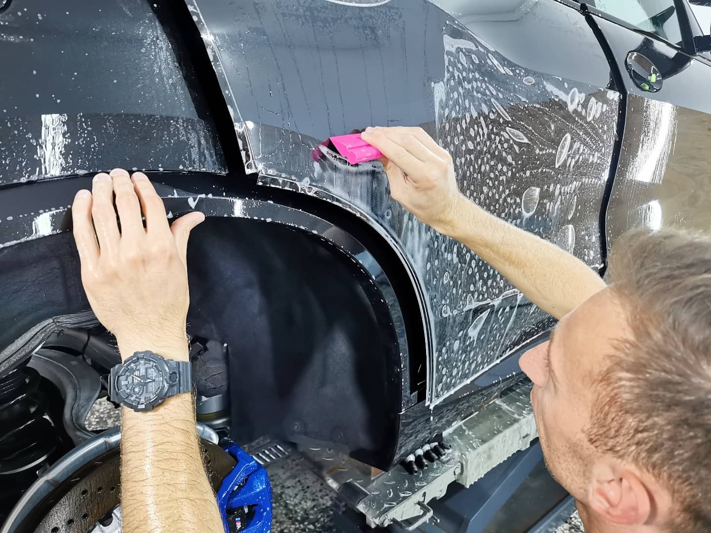 Detailer aplikuje folię ochronną na czarny samochód, używając różowej rakli; widoczny zegarek na nadgarstku