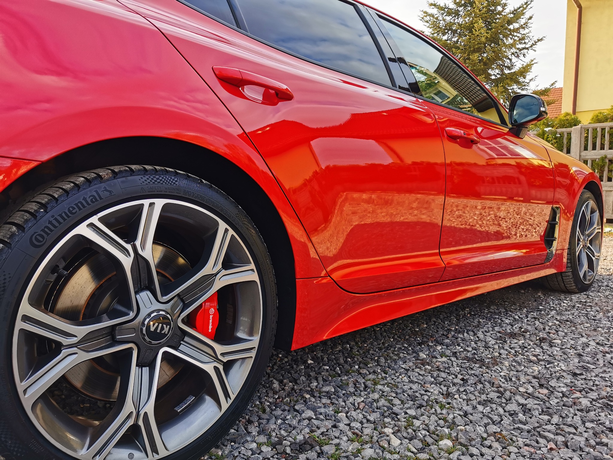 Błyszczący, czerwony samochód Kia Stinger z aluminiowymi felgami i czerwonymi zaciskami hamulcowymi Brembo, sfotografowany z niskiej perspektywy na żwirowym podłożu.