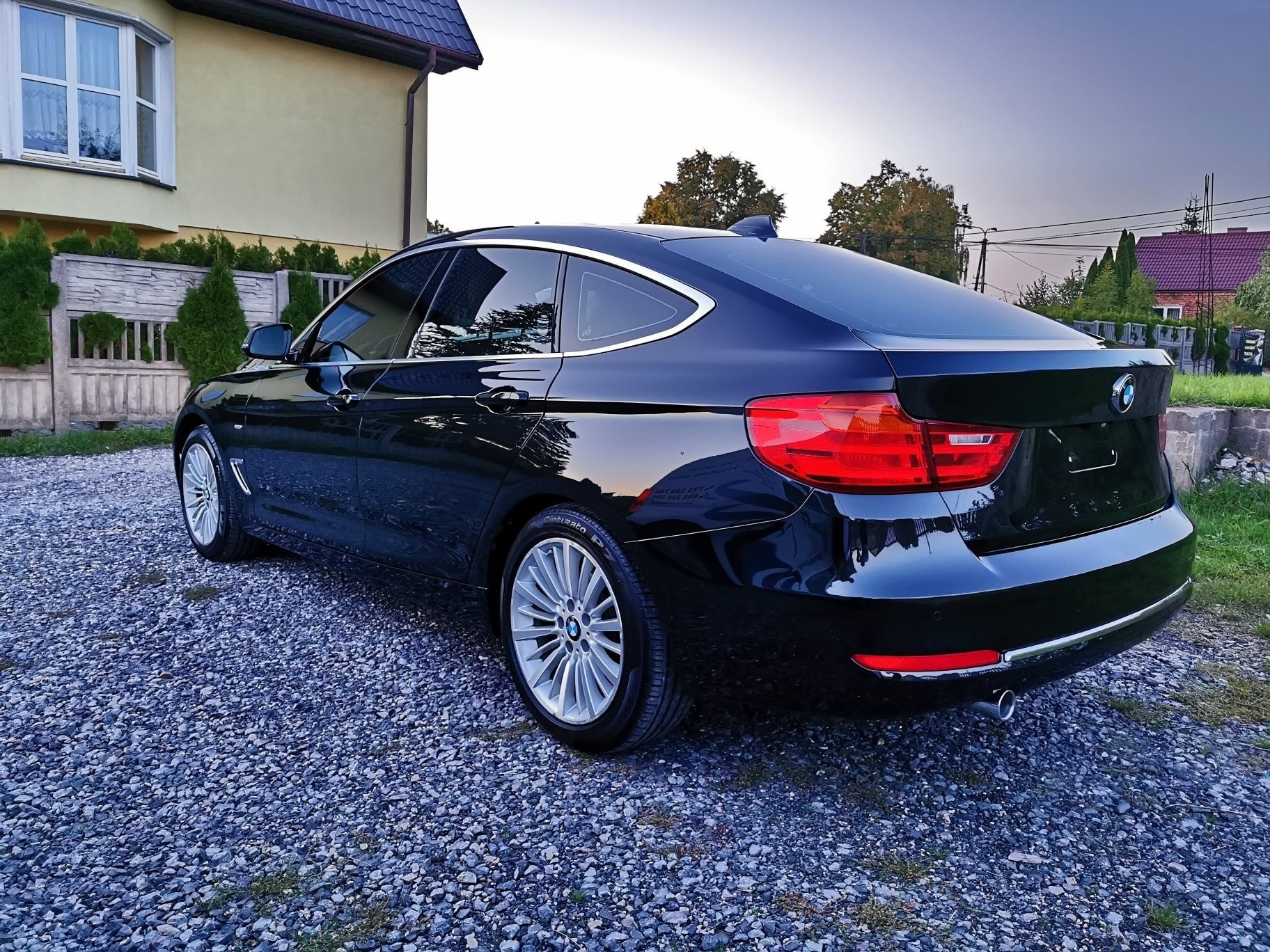 Błyszczący, czarny samochód marki BMW zaparkowany na żwirowym podjeździe przed domem, widok z tyłu i boku, oświetlony popołudniowym słońcem.
