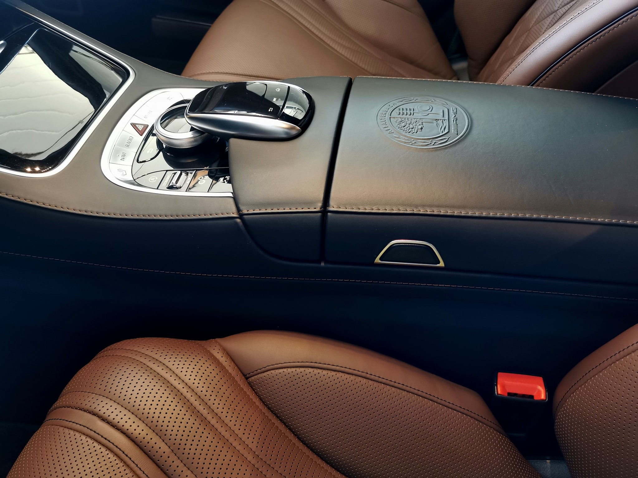 Wnętrze luksusowego samochodu z brązową skórzaną tapicerką, widoczny panel sterowania z chromowanymi elementami i logo producenta na podłokietniku.