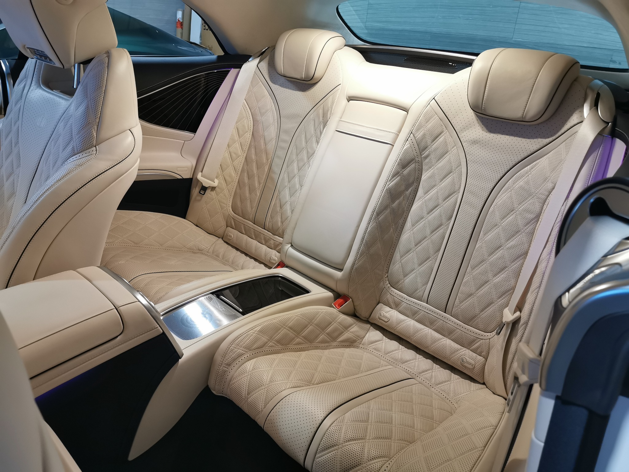 Jasna, skórzana tapicerka tylnych siedzeń luksusowego samochodu z perforacją i pikowaniem w romby, widoczne pasy bezpieczeństwa i podłokietnik z elementami metalowymi.