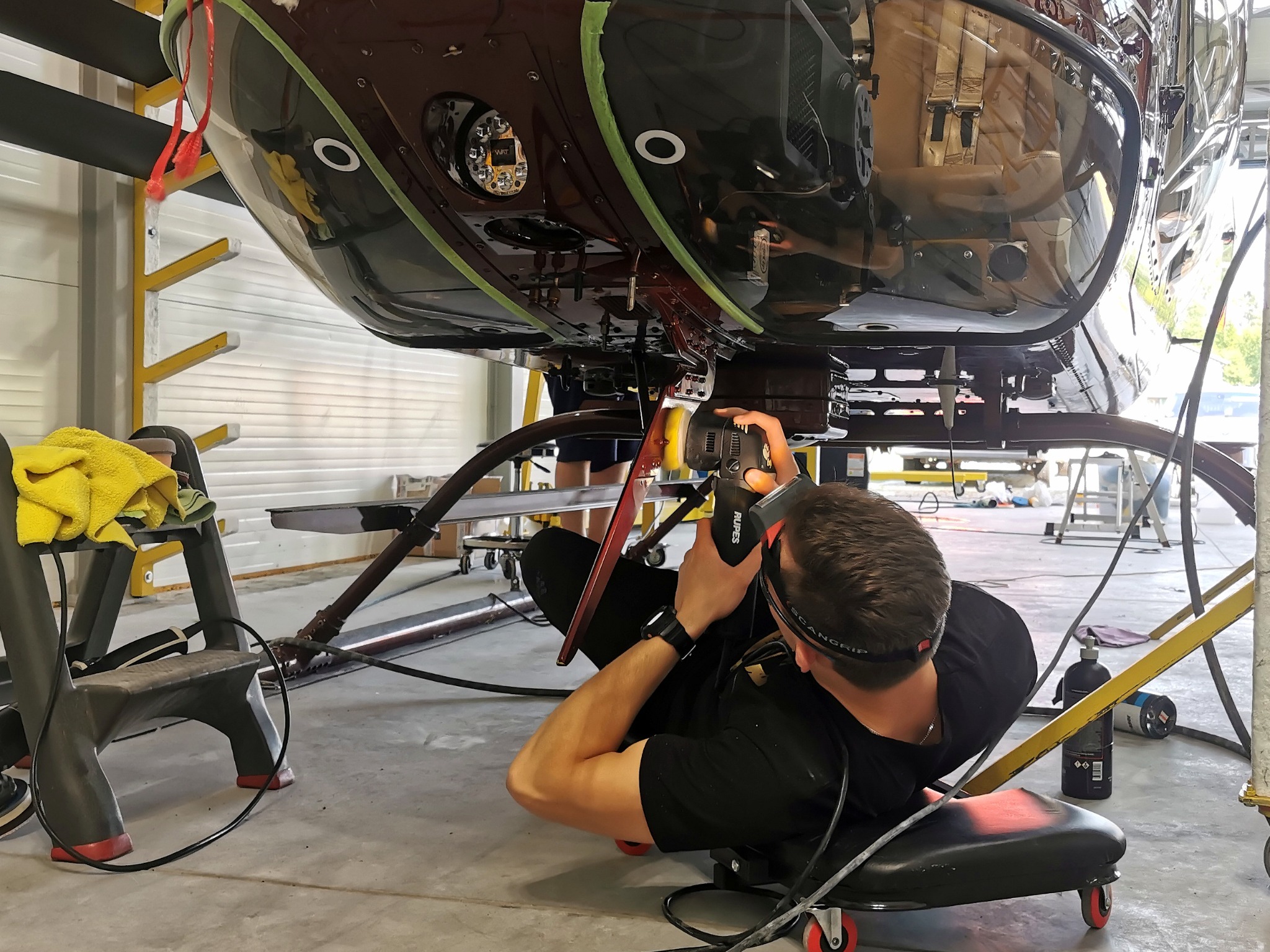 Polerowanie lakieru na podwoziu helikoptera przez mężczyznę w warsztacie, używającego polerki elektrycznej i leżącego na wózku warsztatowym