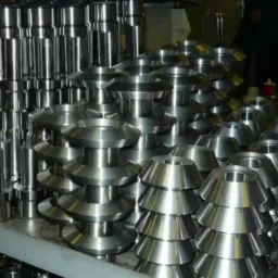 Stalowe, obrobione elementy o różnych kształtach ułożone na półce, widoczne cylindryczne wałki i stożkowe tuleje, metaliczny połysk, w tle regał warsztatowy.