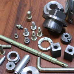 Zestaw różnorodnych metalowych elementów obrobionych na tokarce i CNC, leżących na drewnianym stole; widoczne śruby, nakrętki, tuleje, pręty i inne detale.