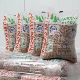 Ułożone na palecie worki z granulatem opałowym, z widocznym logo producenta 'Tartak Olczyk' i certyfikatem DIN Plus, na tle białej ściany.